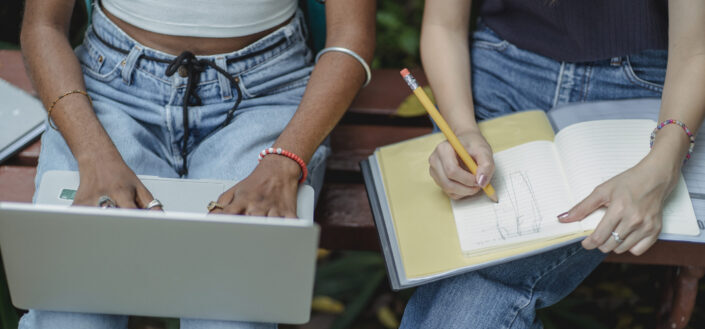 2 women sitting beside each other, one holding a laptop, the other holding a notebook