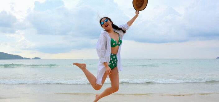 Woman Jumping on Seashore and Holding Hat